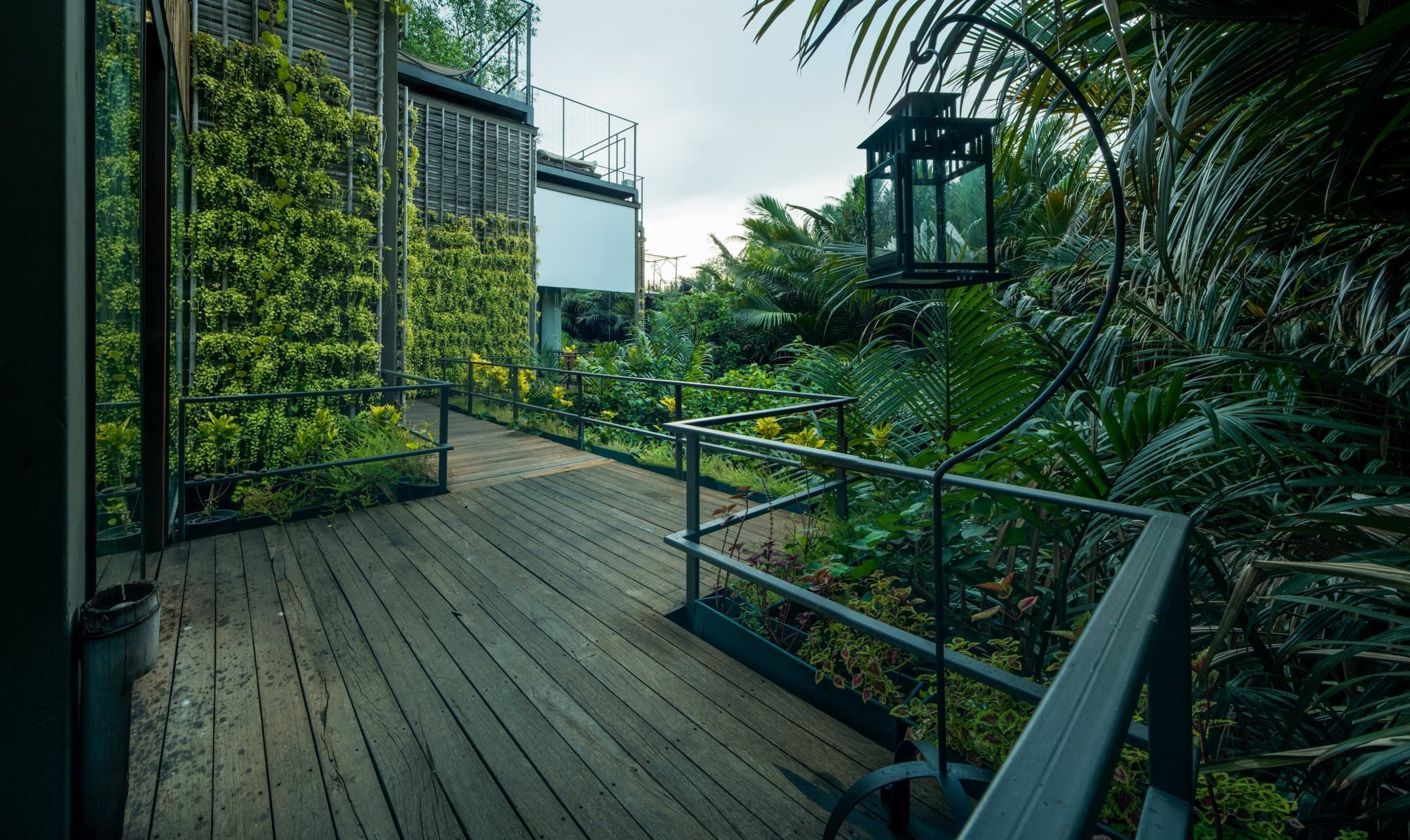 A wooden deck with metal railings, surrounded by lush greenery and a modern building with vertical gardens.