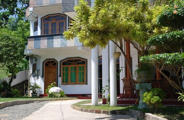 A two-story building with a central entrance, surrounded by a landscaped garden with various plants and a pathway leading to the entrance. - Castle Bay Resort (Weligama)
