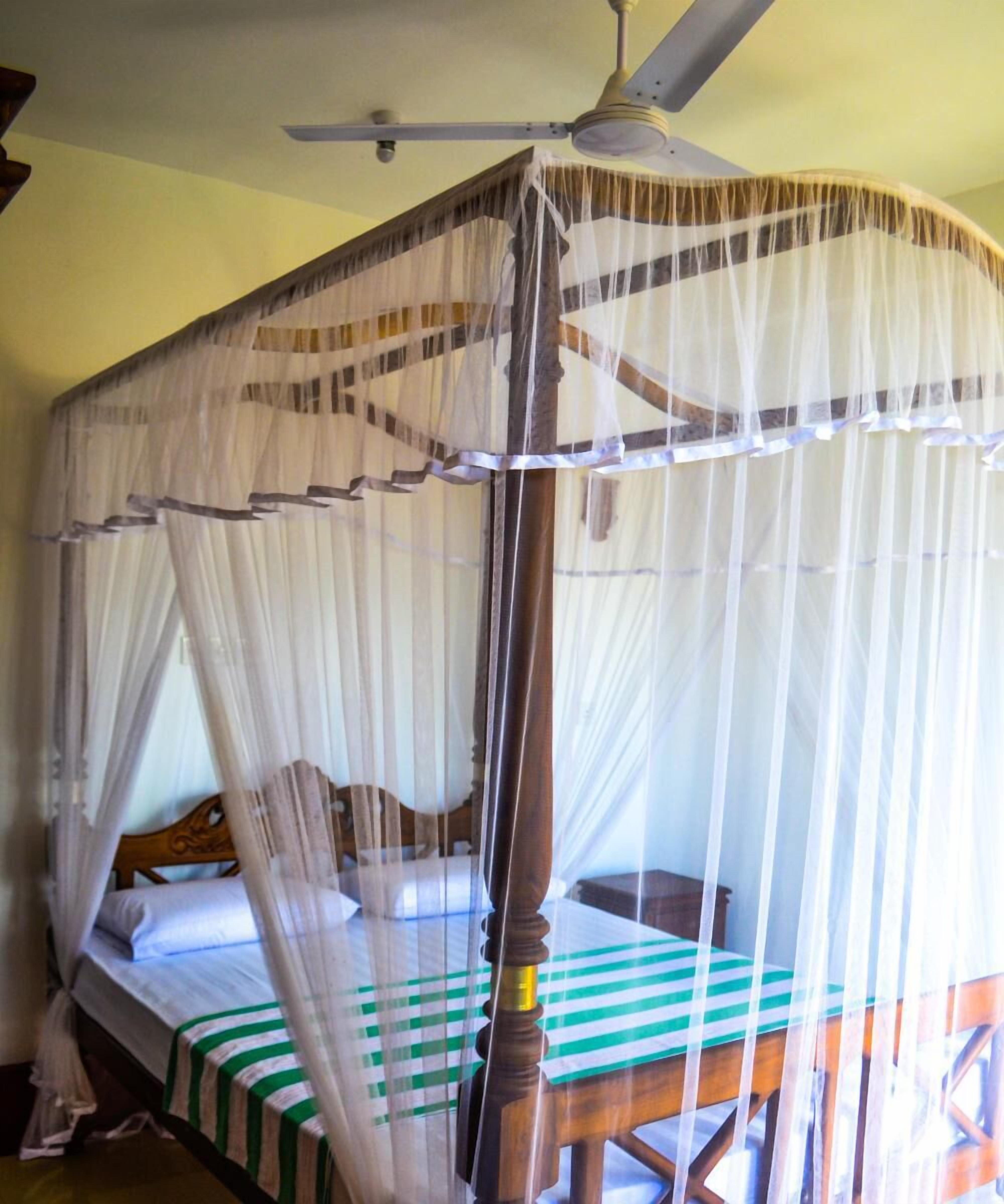 A room with a bed under a mosquito net, a wooden chair, and a ceiling fan.