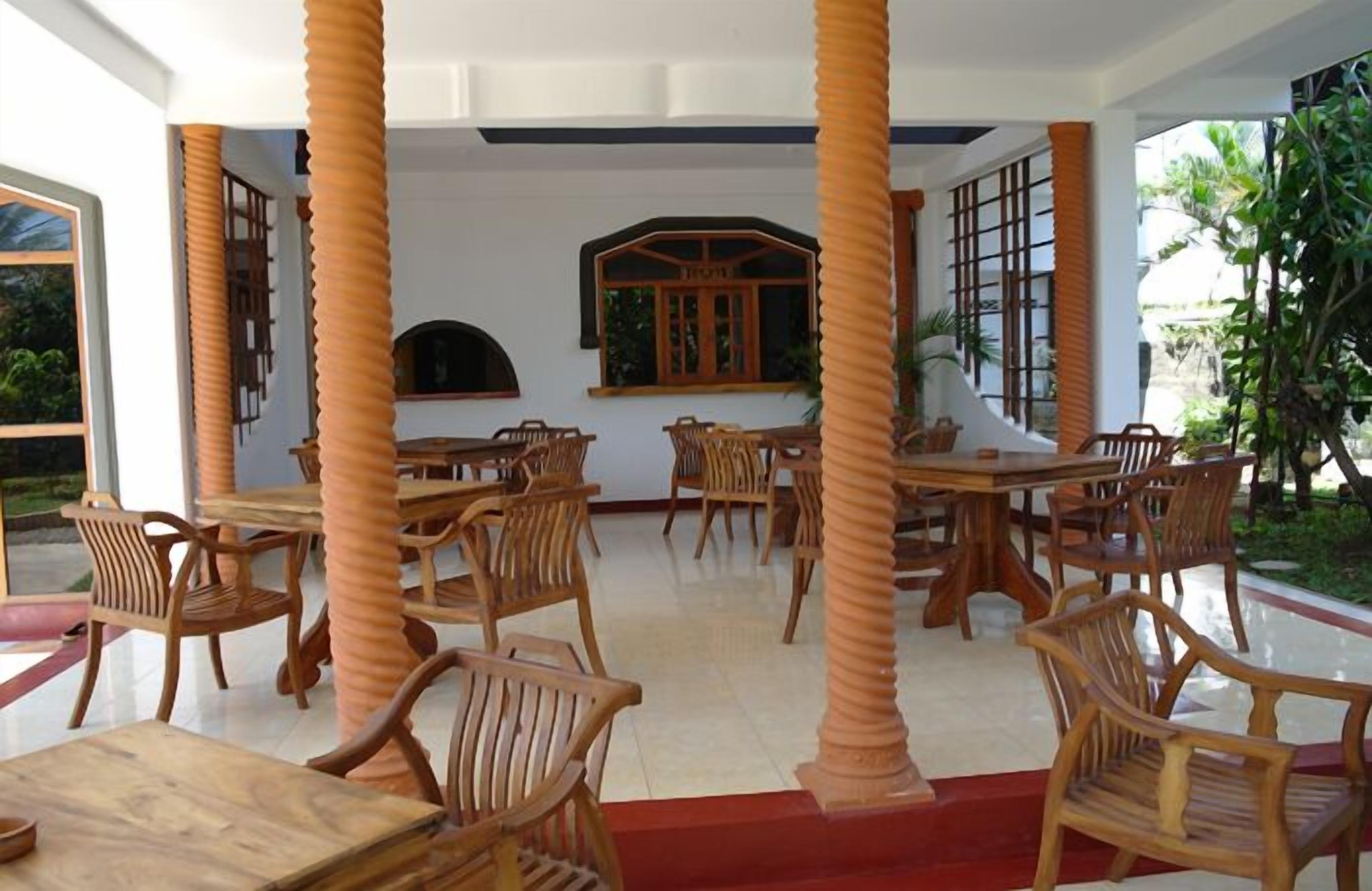 Un restaurante con mobiliario de madera y columnas, que presenta un suelo de baldosas y amplios ventanales.