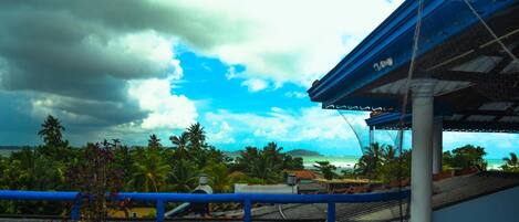 Vista costera con una estructura de techo azul, palmeras y cielo nublado.