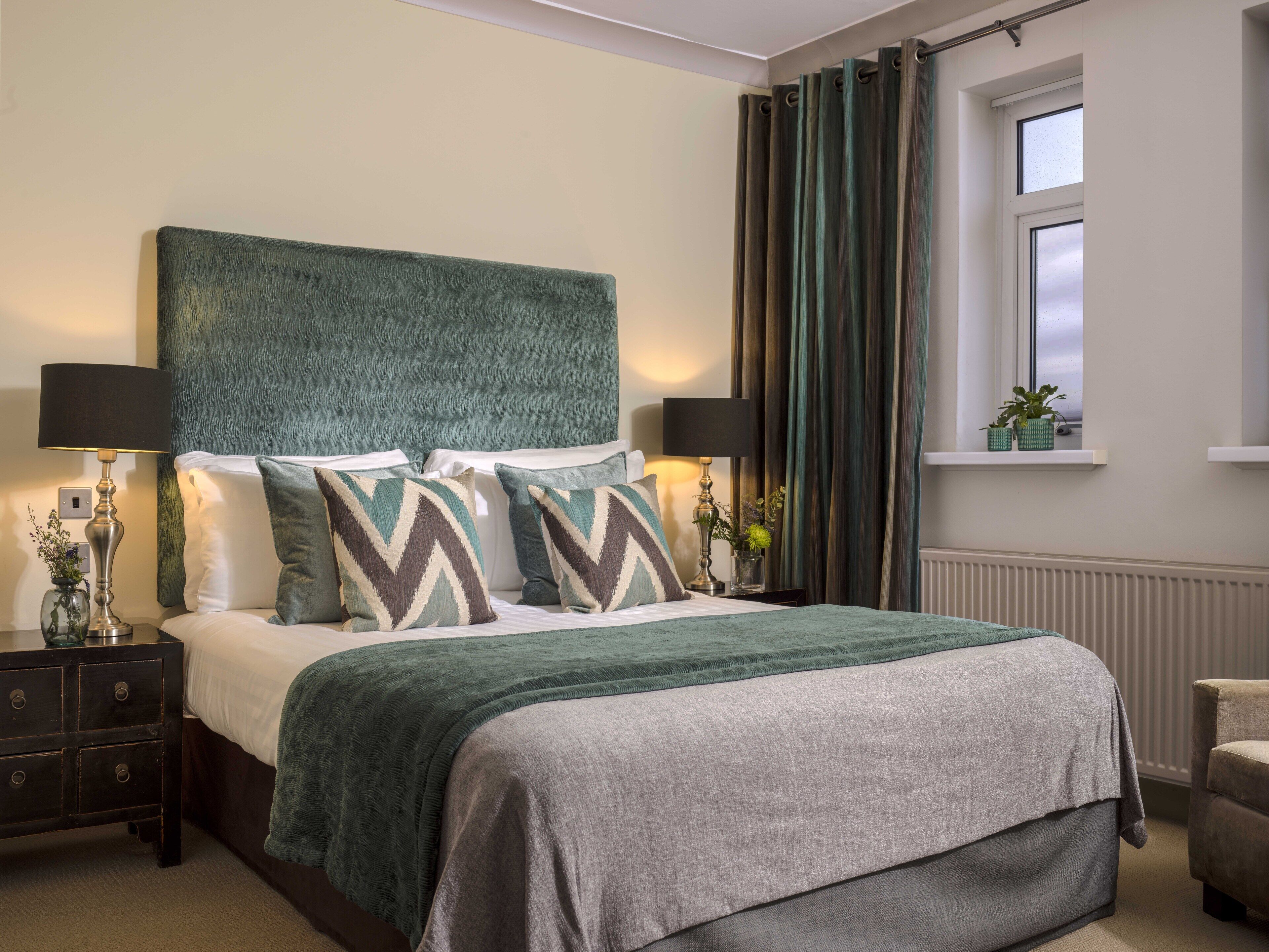 A neatly arranged bedroom with a large bed, two bedside tables with lamps, a window with curtains, and a small plant on the windowsill.