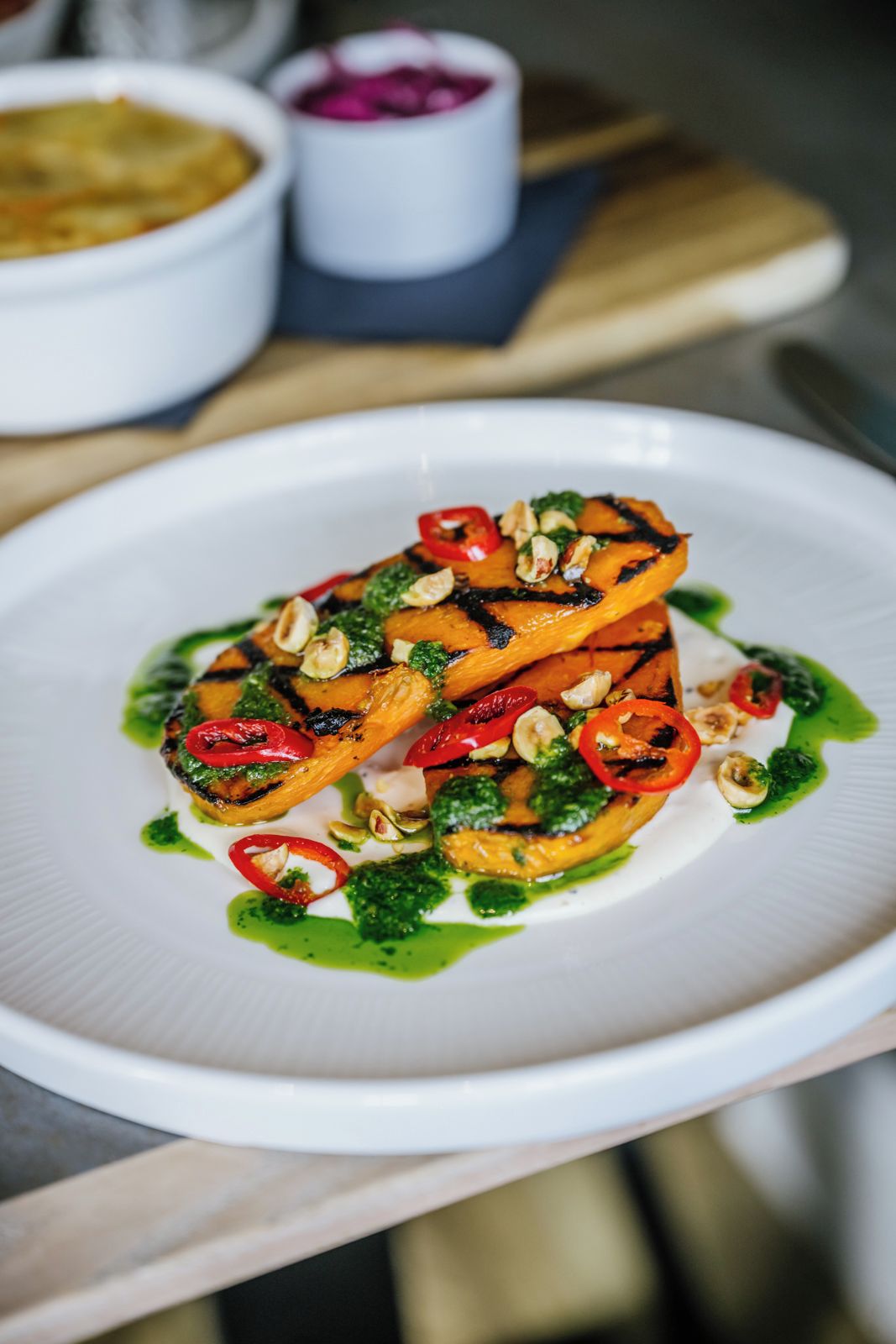 A white plate with a dish of grilled vegetables and nuts, accompanied by a green sauce.