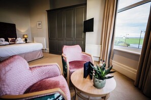 A hotel room with a bed, two pink armchairs, a small table with a plant, a TV, and a view of a green landscape through a window. - Clifton Arms Hotel (Lytham St. Anne's)