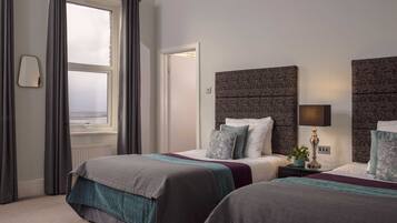 A hotel room with two beds, a large window with curtains, a bedside lamp, and a view of the sea.