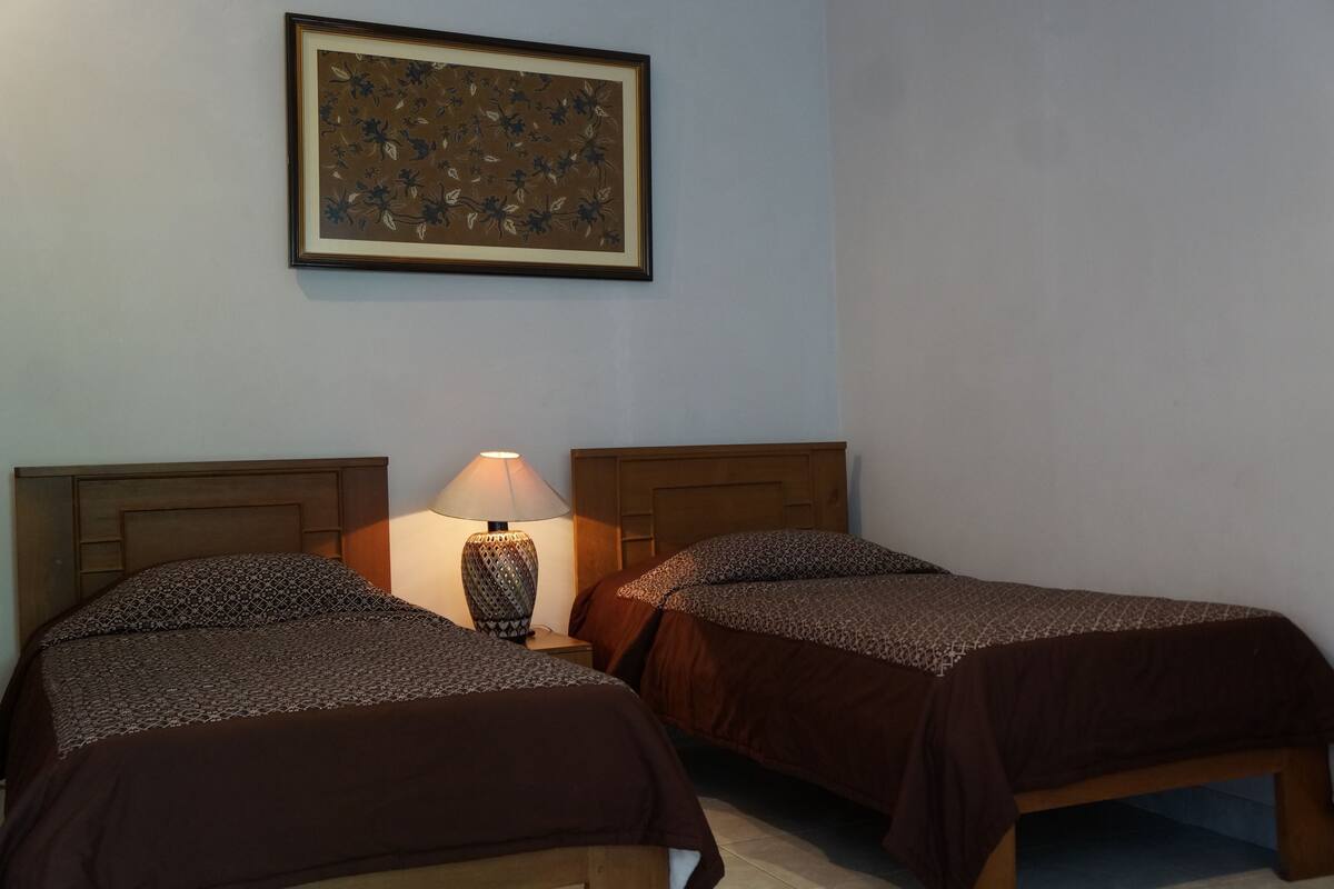 Two beds with patterned bedding, a nightstand with a lamp, and a framed floral artwork on the wall.