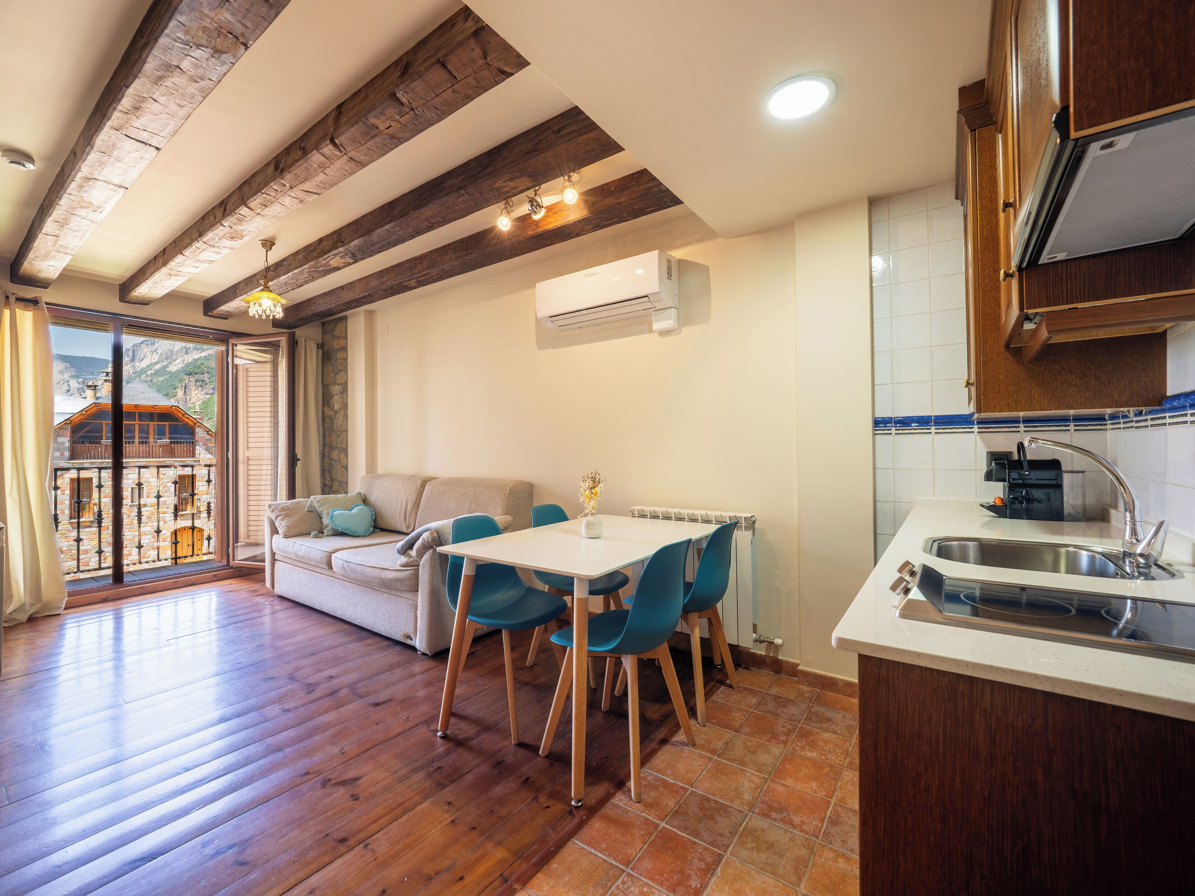 Una cucina accogliente con travi in legno, un'area pranzo con sedie blu e vista su un edificio in pietra attraverso una porta in vetro.