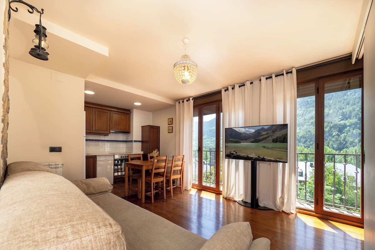 A living room with a sofa, a dining area with a table and chairs, a kitchen with cabinets and appliances, and a balcony with a view of the mountains.