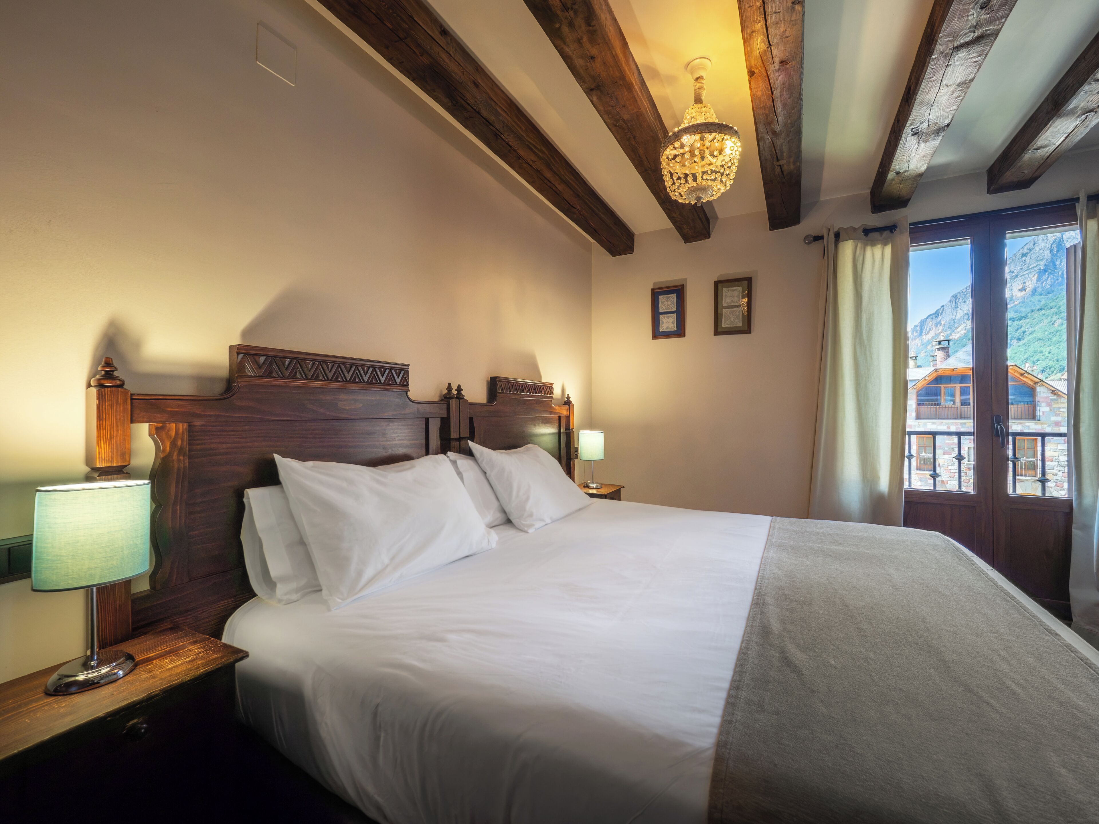 Habitación de hotel con una cama grande, cabecera de madera, dos lámparas de noche, un candelabro y vistas a la montaña a través de la ventana.