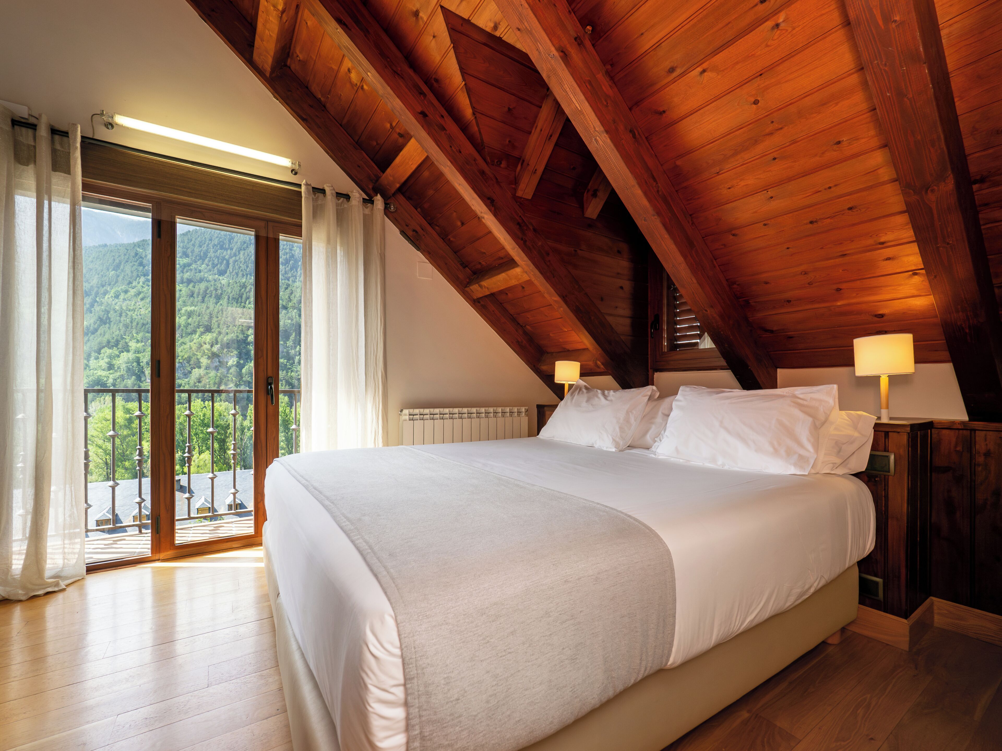A bedroom with a large bed, wooden ceiling beams, a view of greenery outside, and a balcony.