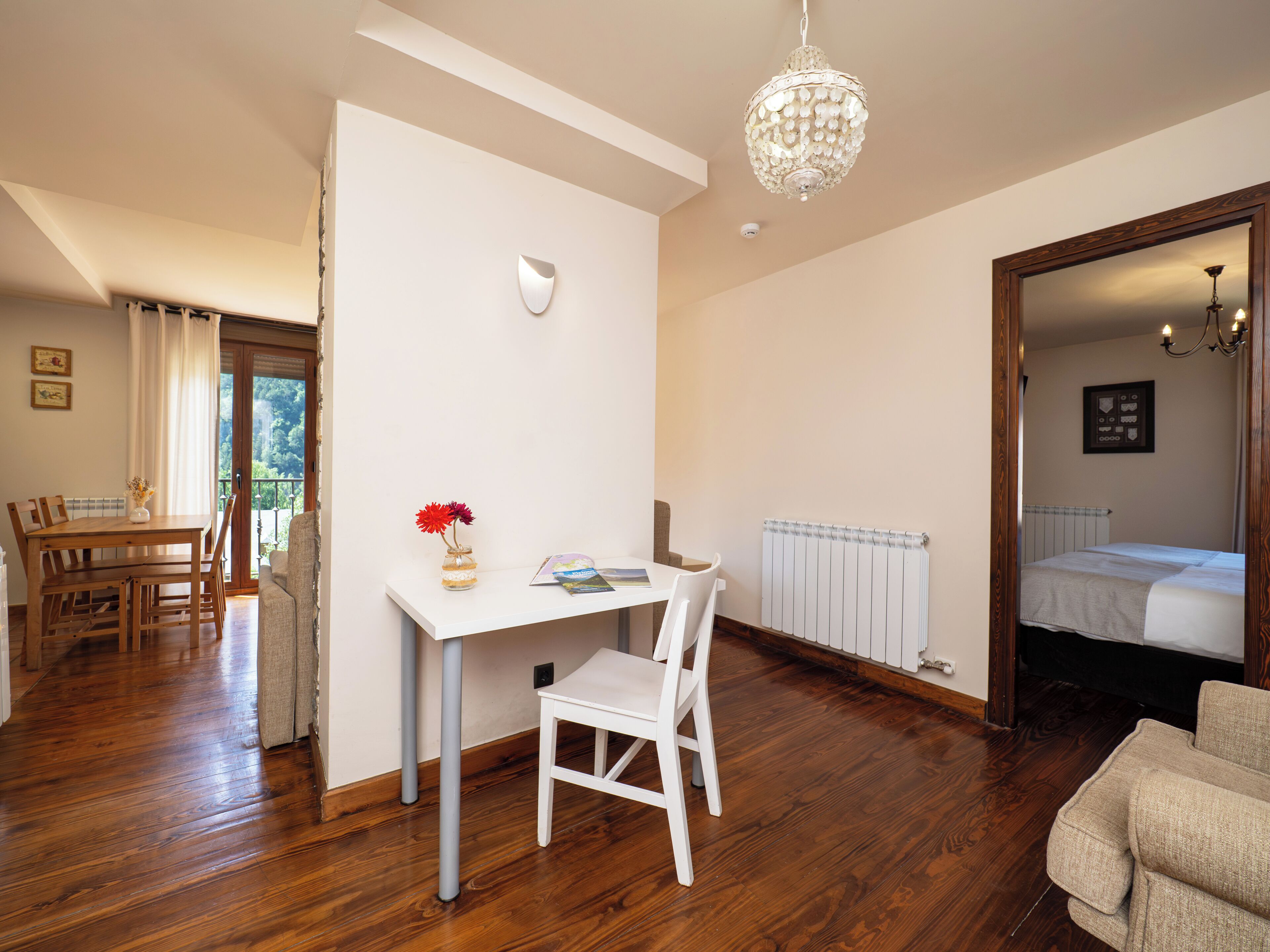 Una habitación con suelo de madera, un escritorio blanco, una silla blanca, un radiador y una cama con cabecero de madera.