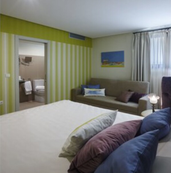 Una habitación de hotel con una cama, un sofá, un cuadro en la pared y un baño visible a través de una puerta abierta.