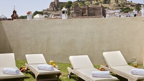 Una terraza en la azotea con cinco sillones de descanso, cada uno con una toalla y un vaso de jugo de naranja. La terraza ofrece vistas a un promontorio histórico con edificios antiguos.