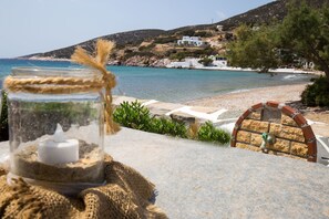 A table with a glass jar containing a candle and a rustic cloth. In the background, there is a beach with white buildings and a clear blue sea. - Pension Ageliki (Sifnos)