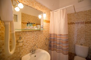 A bathroom with a glass shelf holding toiletries, a white sink, a shower with a striped curtain, and a tiled wall. - Pension Ageliki (Sifnos)