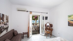 A room with a sofa, a bed, a desk with a chair, and a view through an open door to an outdoor area with a table and chairs. - Pension Ageliki (Sifnos)
