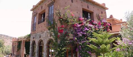 A stone building with a balcony, surrounded by lush greenery and flowering plants.