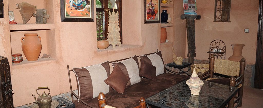 A cozy room with a brown sofa, a small table, and shelves with pottery and decorative items.