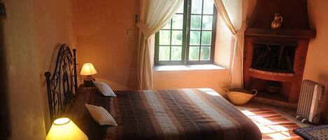 A bedroom with a bed, two bedside lamps, a window with curtains, a fireplace, and a radiator.