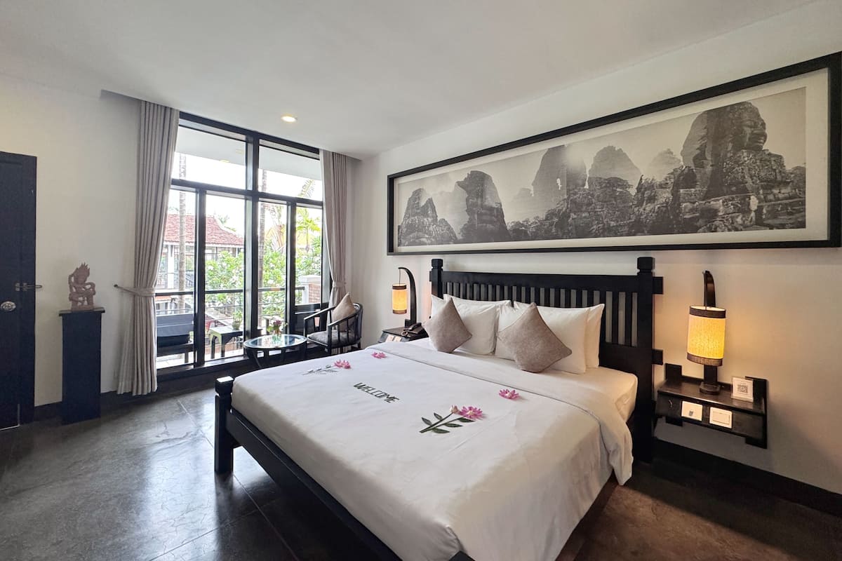 A hotel room with a large bed, a nightstand with a lamp, a chair, a window with curtains, and a framed picture on the wall.