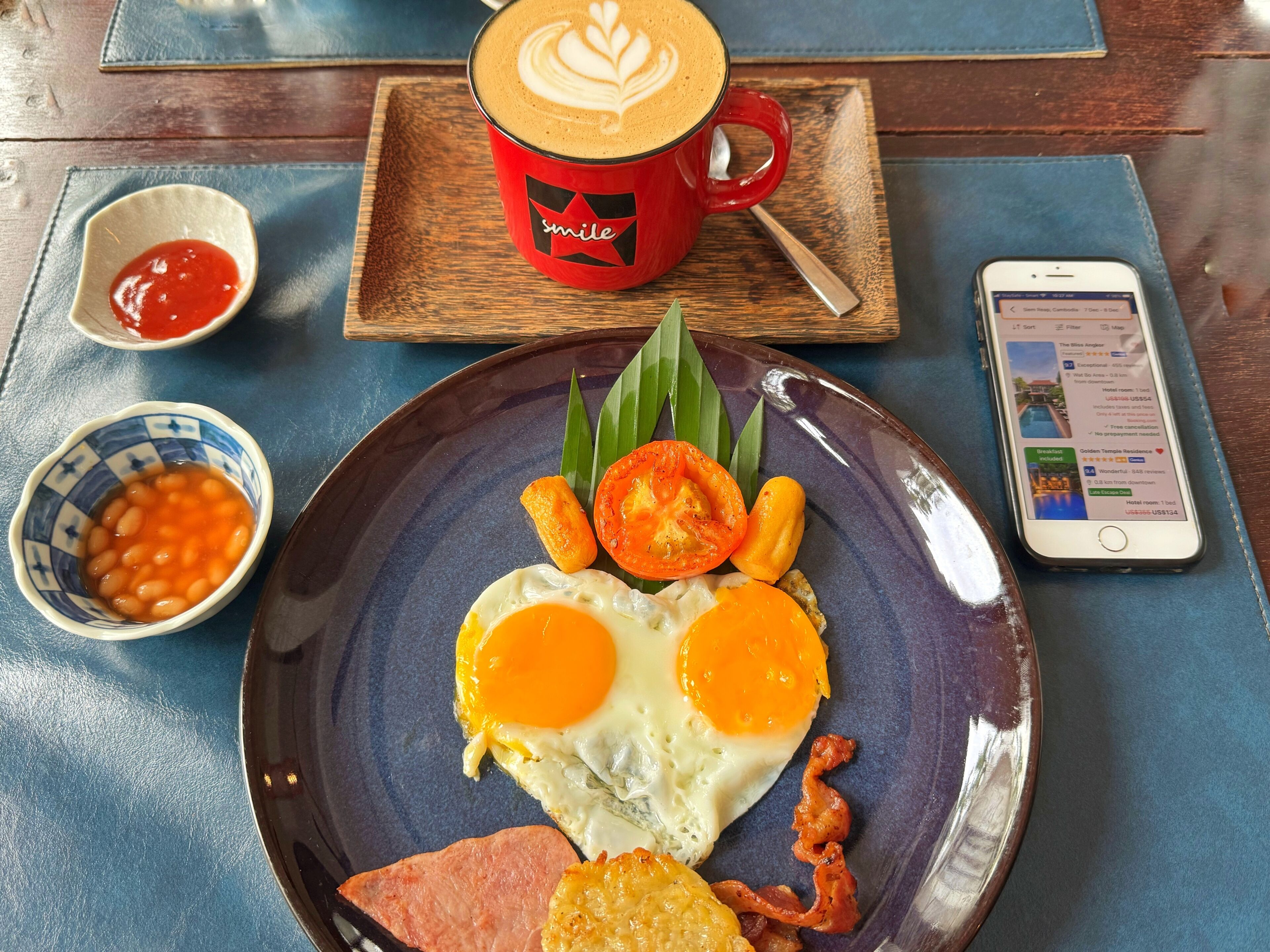 Un plat d'esmorzar amb ous, bacon i fesols, una tassa vermella amb un dibuix de làtex, un telèfon intel·ligent que mostra una reserva d'hotel, i una safata de fusta.