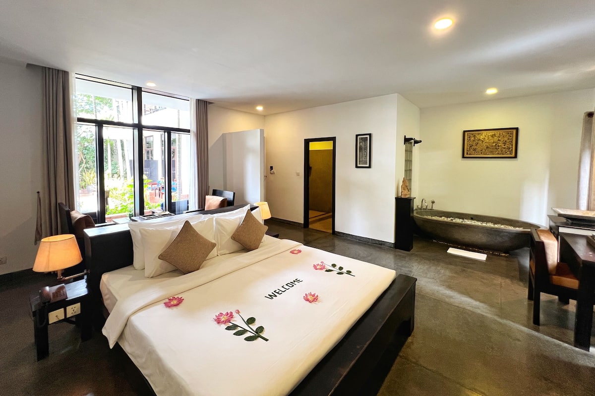 A hotel room with a large bed, a sitting area with a sofa and armchairs, a bathtub, and a view of the outdoors through large windows.