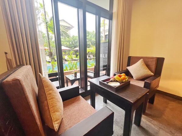 Una habitación con dos sillones acolchados, una mesita con fruteros y vistas a una piscina y follaje exterior.