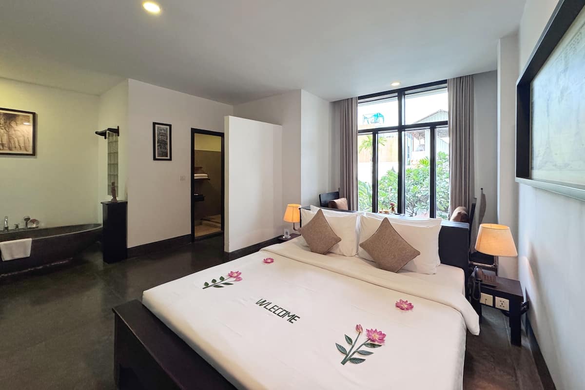 A hotel room with a bed, bedside lamps, a window with a view, a bathroom with a bathtub, and a door leading to another room.