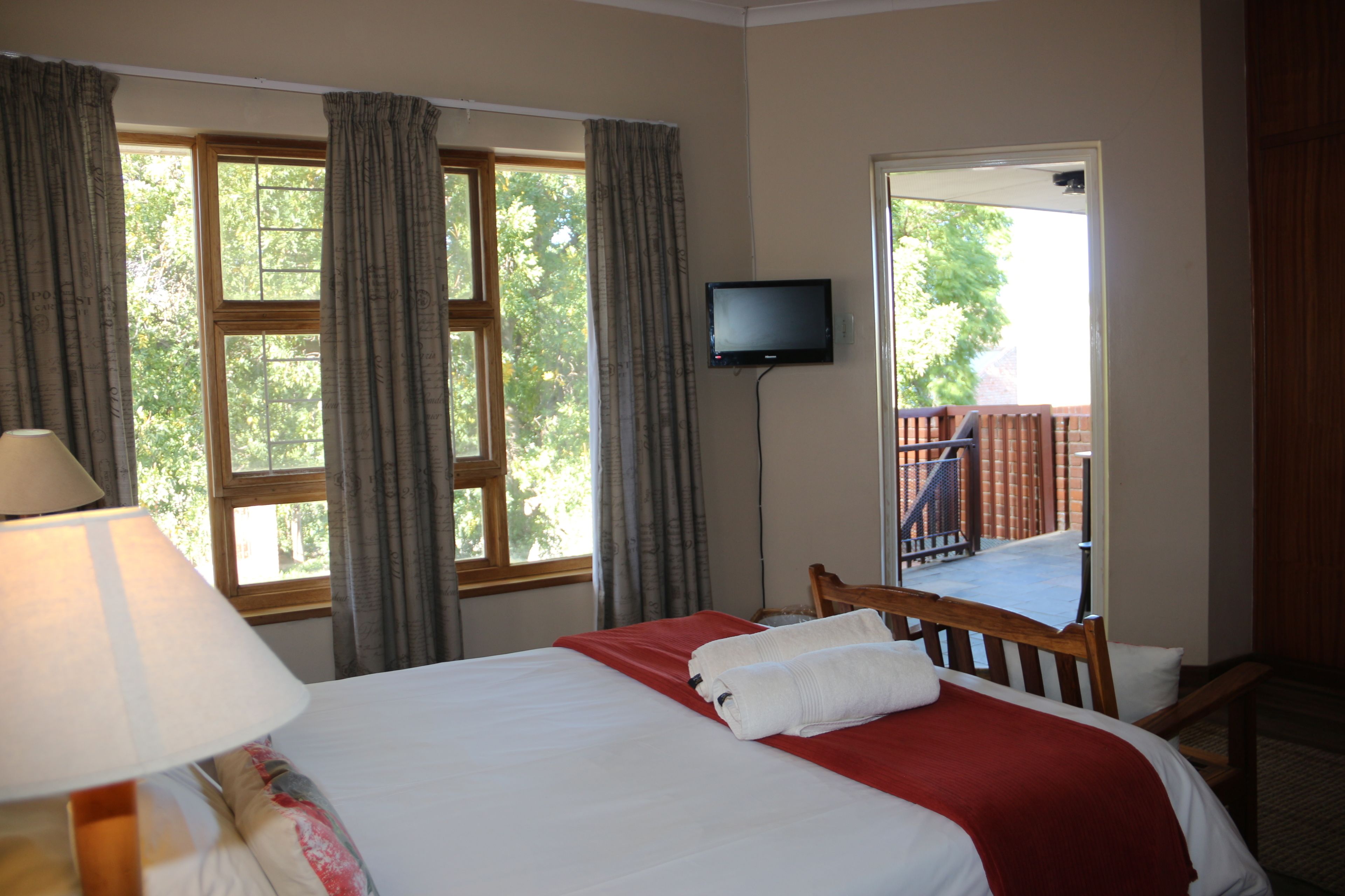 A hotel room with a bed, a TV, a window with curtains, and a view of an outdoor deck.