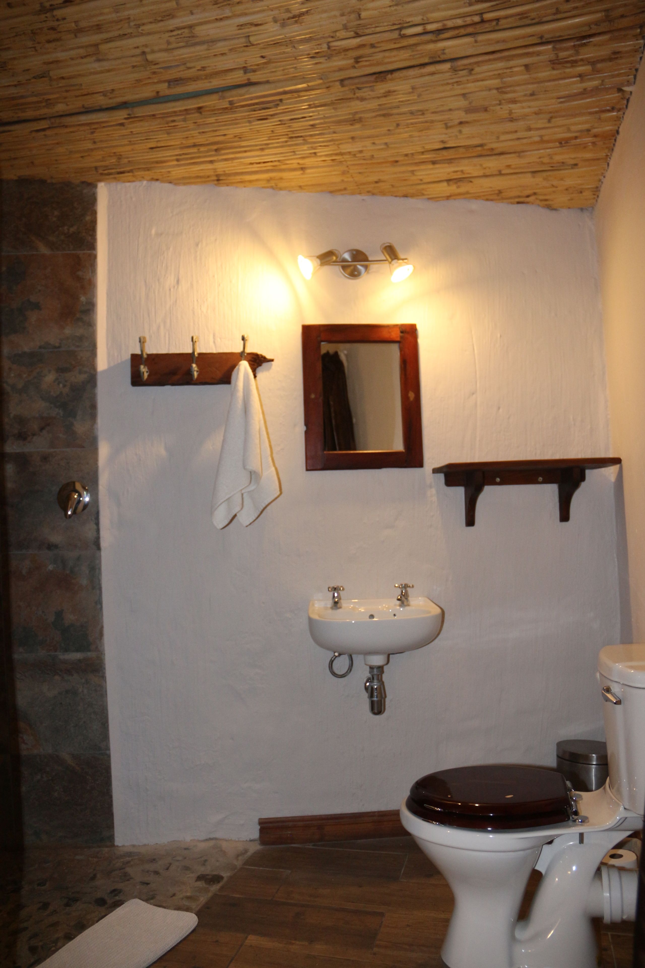 Casa de banho com parede de pedra, pia branca, sanita de madeira e espelho sobre a pia.