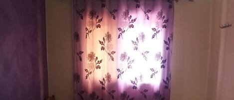Un dormitorio con cama, ropa de cama morada y azul, una cortina floral y un cabecero de madera.