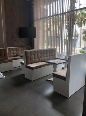 Un área de descanso moderna con asientos acolchados y una mesa de vidrio, ventanas amplias con persianas verticales y un televisor de pantalla plana montado en la pared.