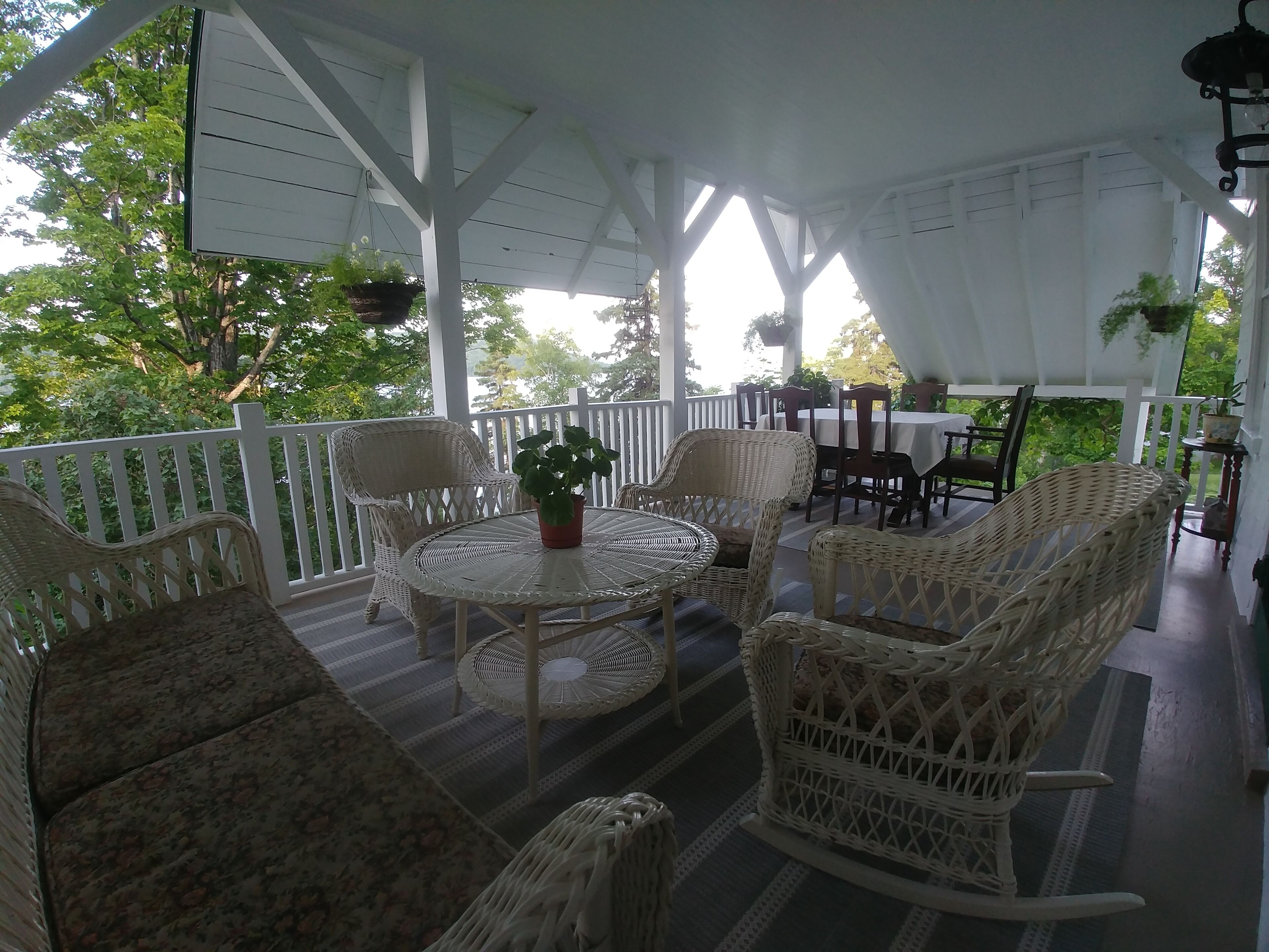 A covered outdoor seating area with wicker furniture and a table, surrounded by white railings and greenery.