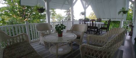 A covered outdoor seating area with wicker furniture and a table, surrounded by white railings and greenery.