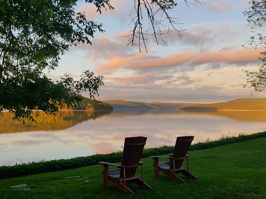 A serene lakeside view with two wooden Adirondack chairs facing the water, surrounded by lush greenery and a clear sky.