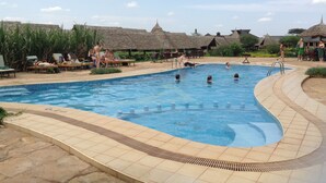 Una piscina con gente en el agua, sillas de descanso y estructuras de techo de paja al fondo.
