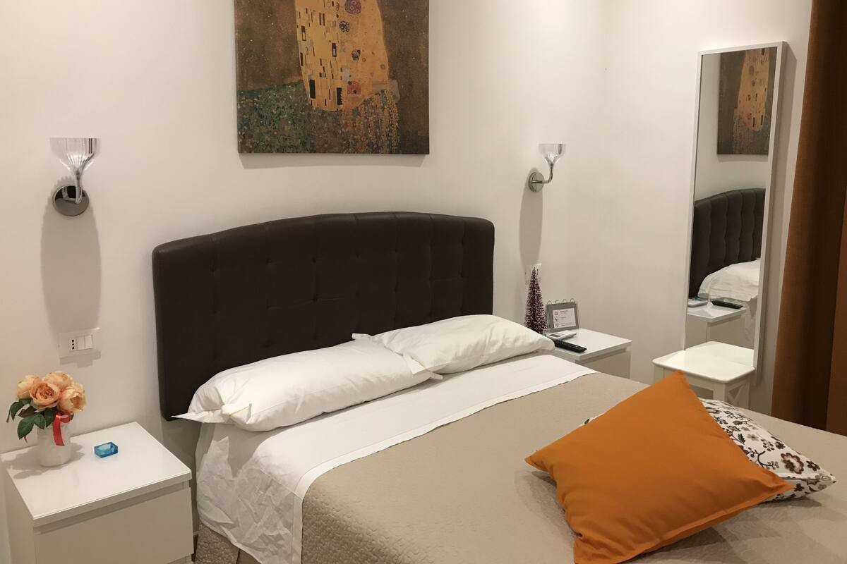 A hotel room with a large bed, white bedding, a dark headboard, a painting above the bed, a bedside table with a vase of flowers, and a nightstand with a lamp.