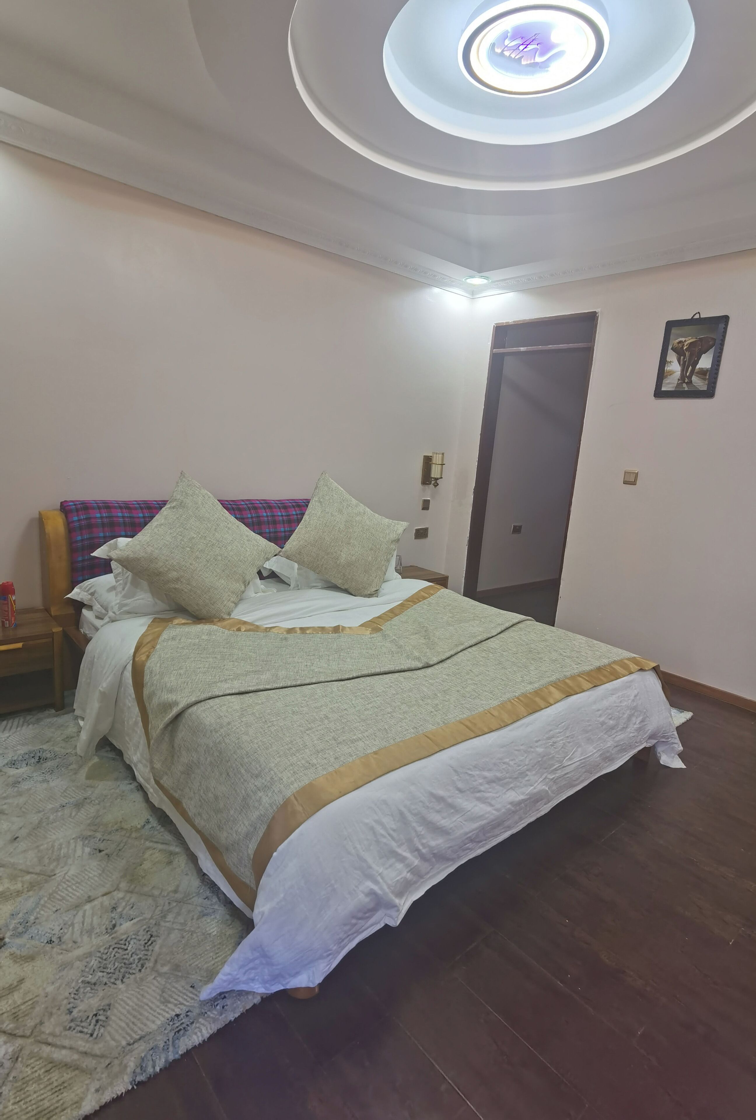 A bedroom with a bed, a nightstand, a wall-mounted light, a door, and a ceiling light fixture.