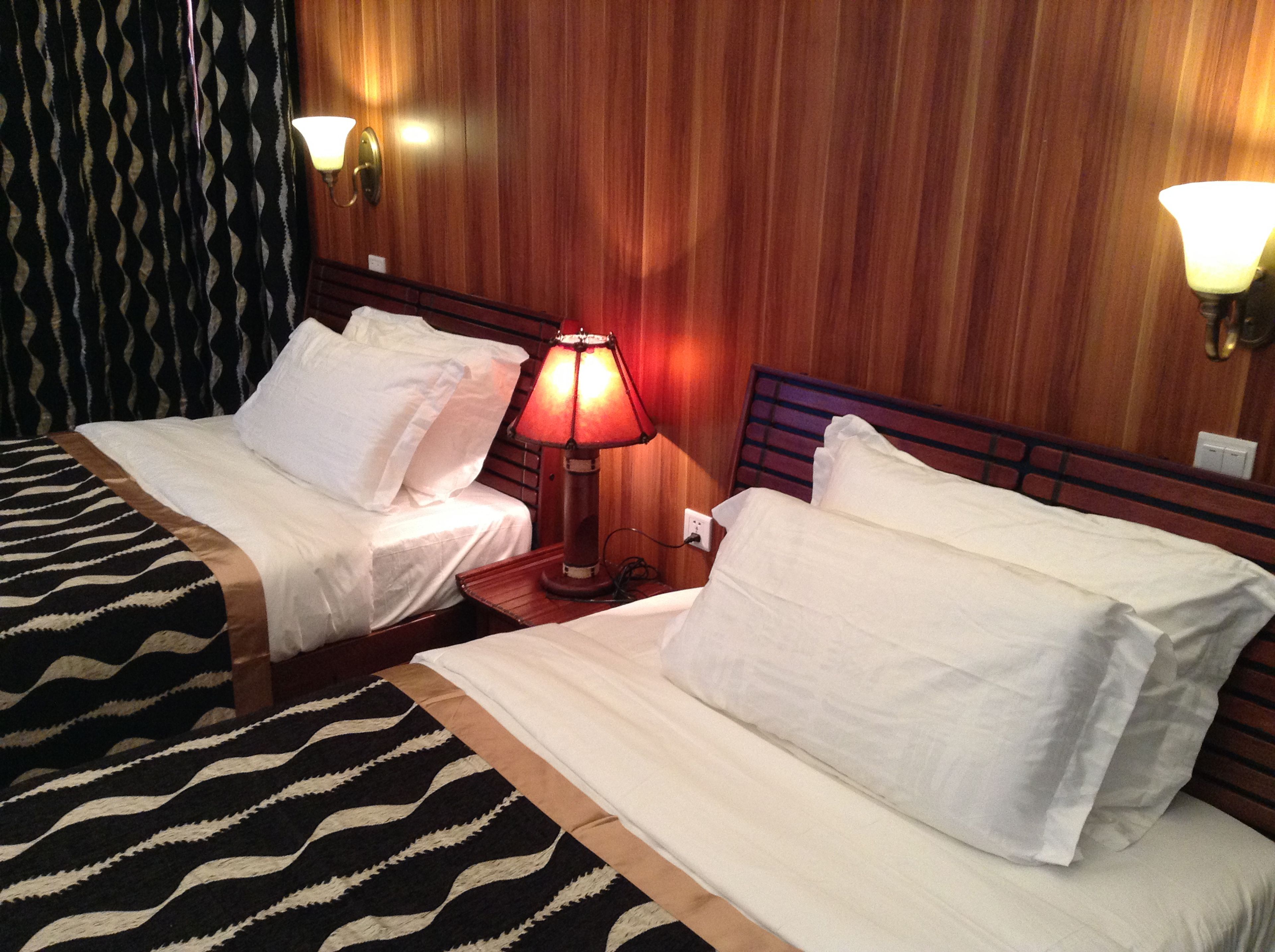 A hotel room with two beds, a patterned rug, wooden walls, and wall-mounted lamps.