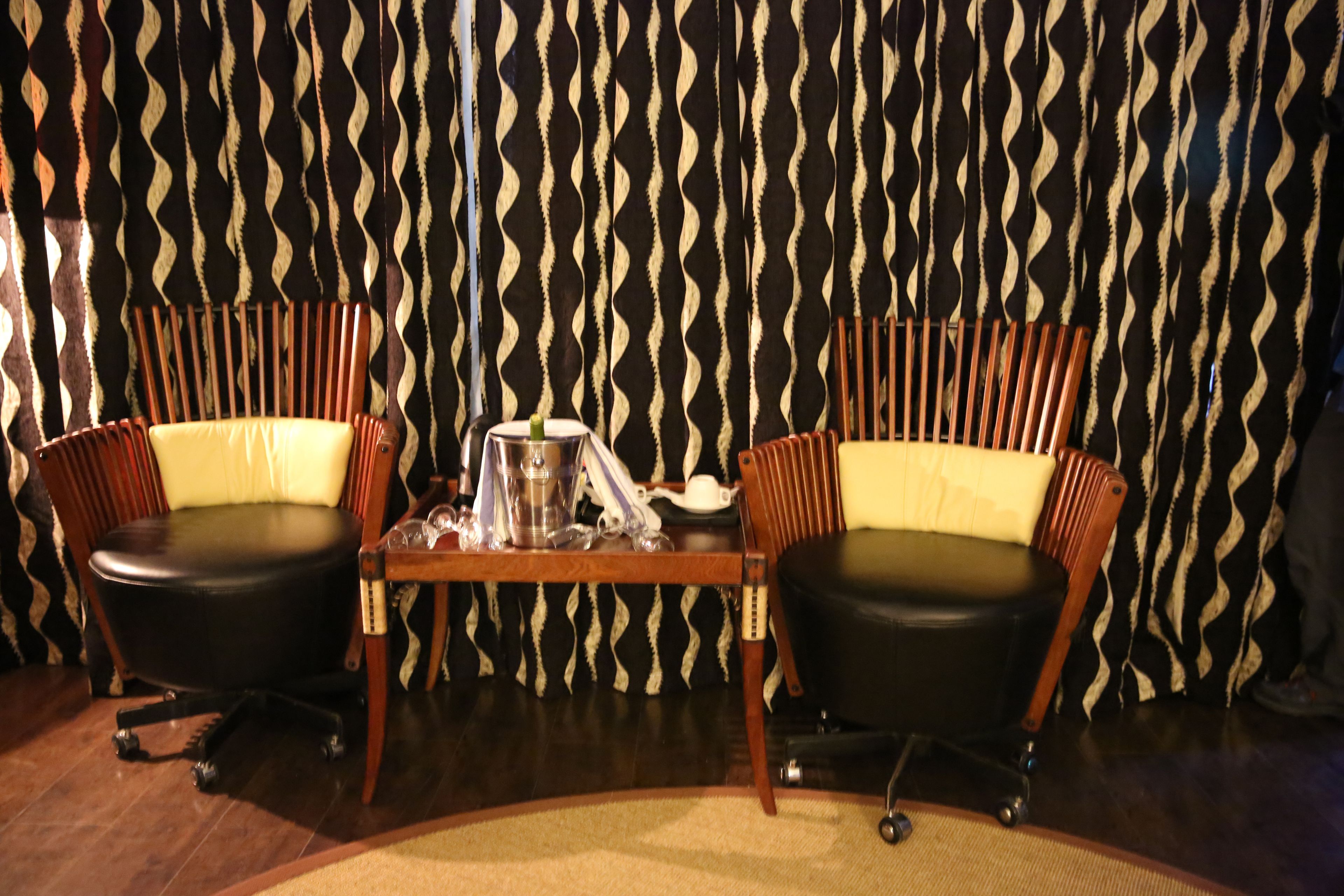 A room with two black leather chairs with wooden frames and yellow cushions, a small wooden table with a metal pitcher and a glass, and a patterned black and white curtain in the background.