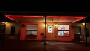 Un edificio con fachada iluminada en rojo, un banco afuera y una puerta con un cartel.