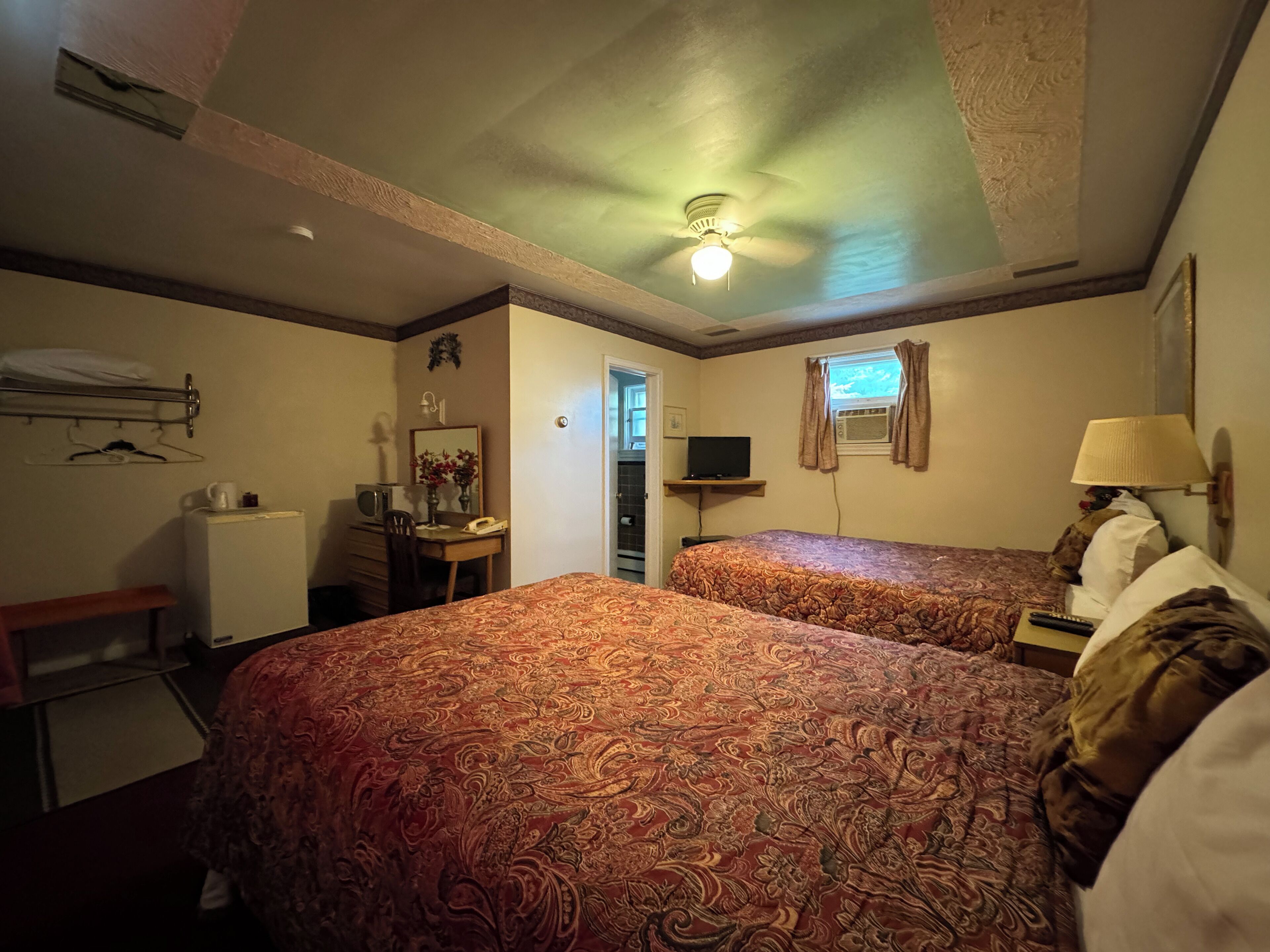 Une chambre d’hôtel avec deux lits, un bureau, une chaise, un miroir, un ventilateur de plafond et une fenêtre avec des rideaux.