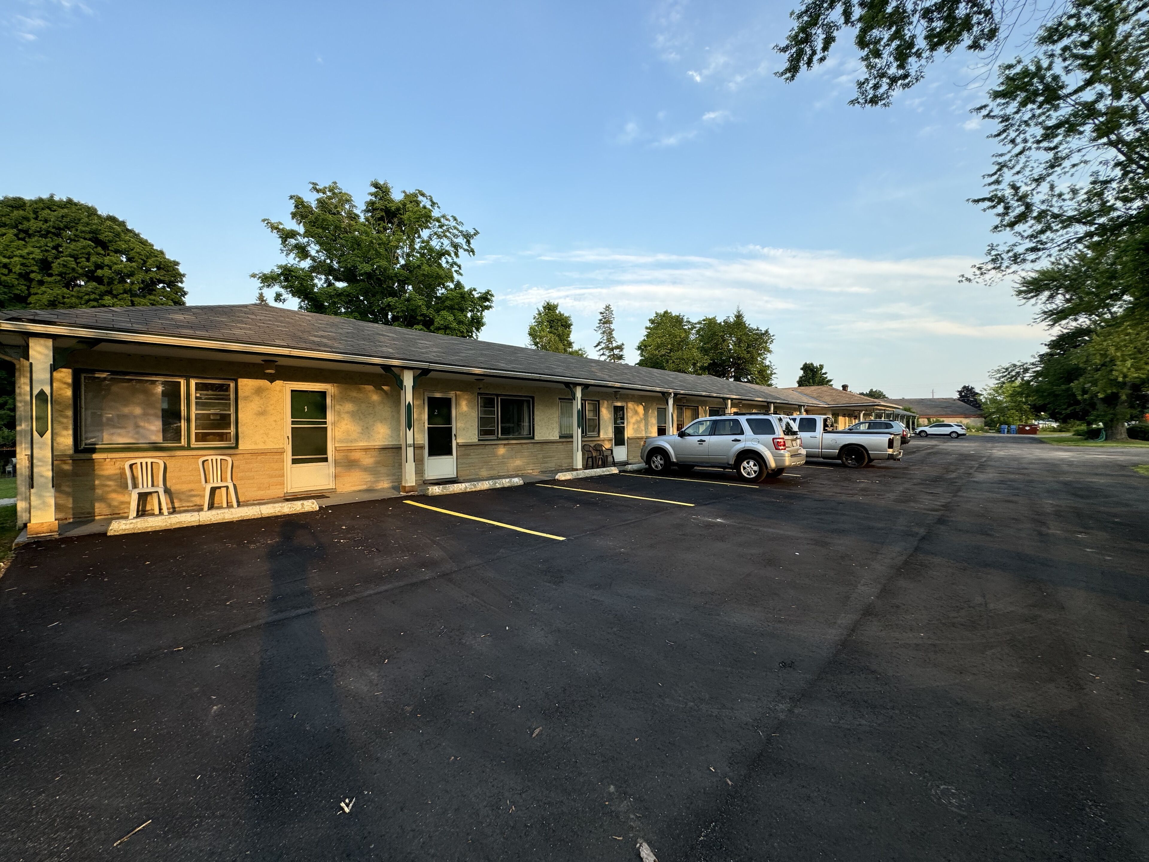 A row of single-story motel buildings with numbered doors, a parking lot, and a few vehicles parked in front.