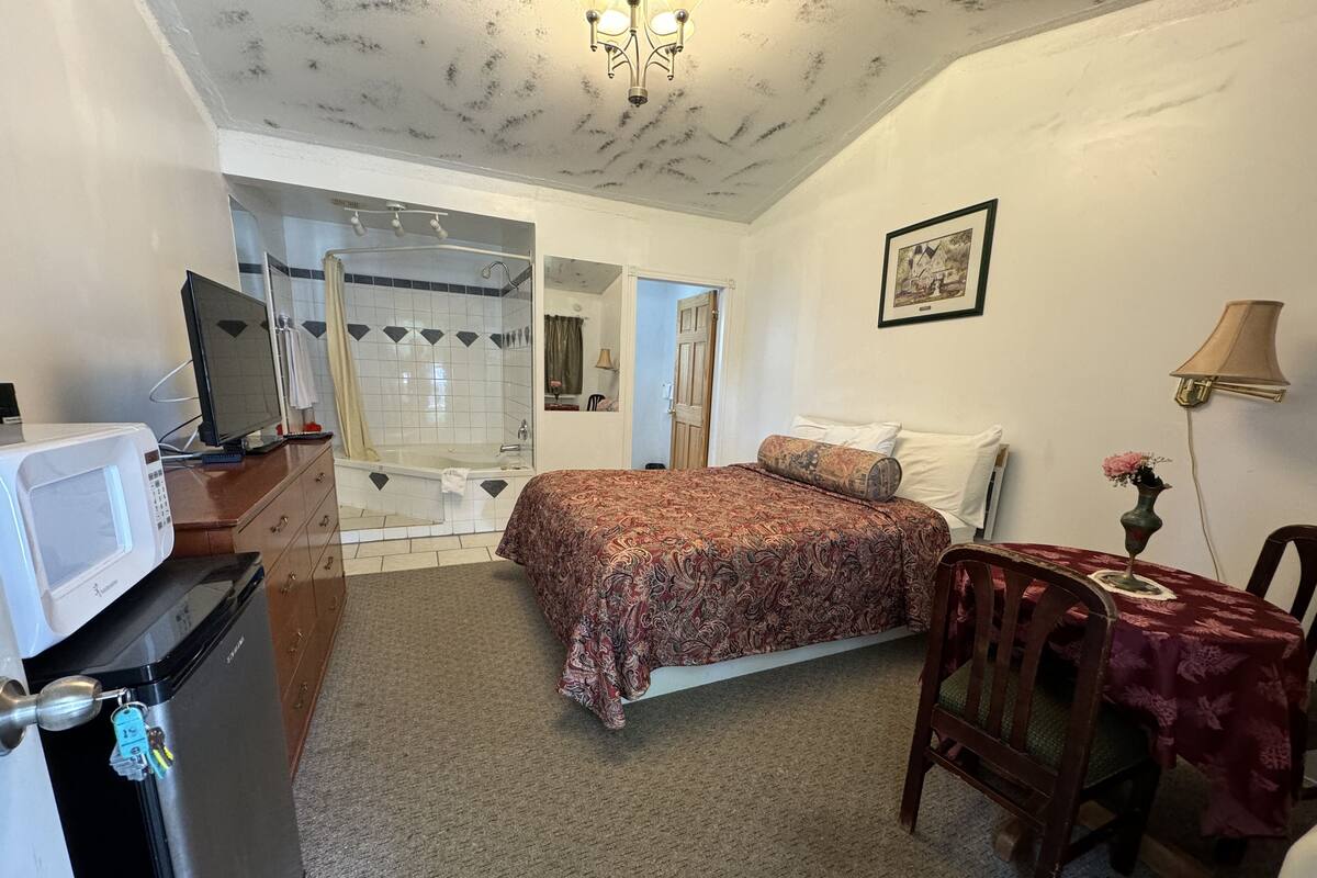 A bedroom with a bed, a desk with a lamp, a microwave, and a bathroom with a bathtub and shower.