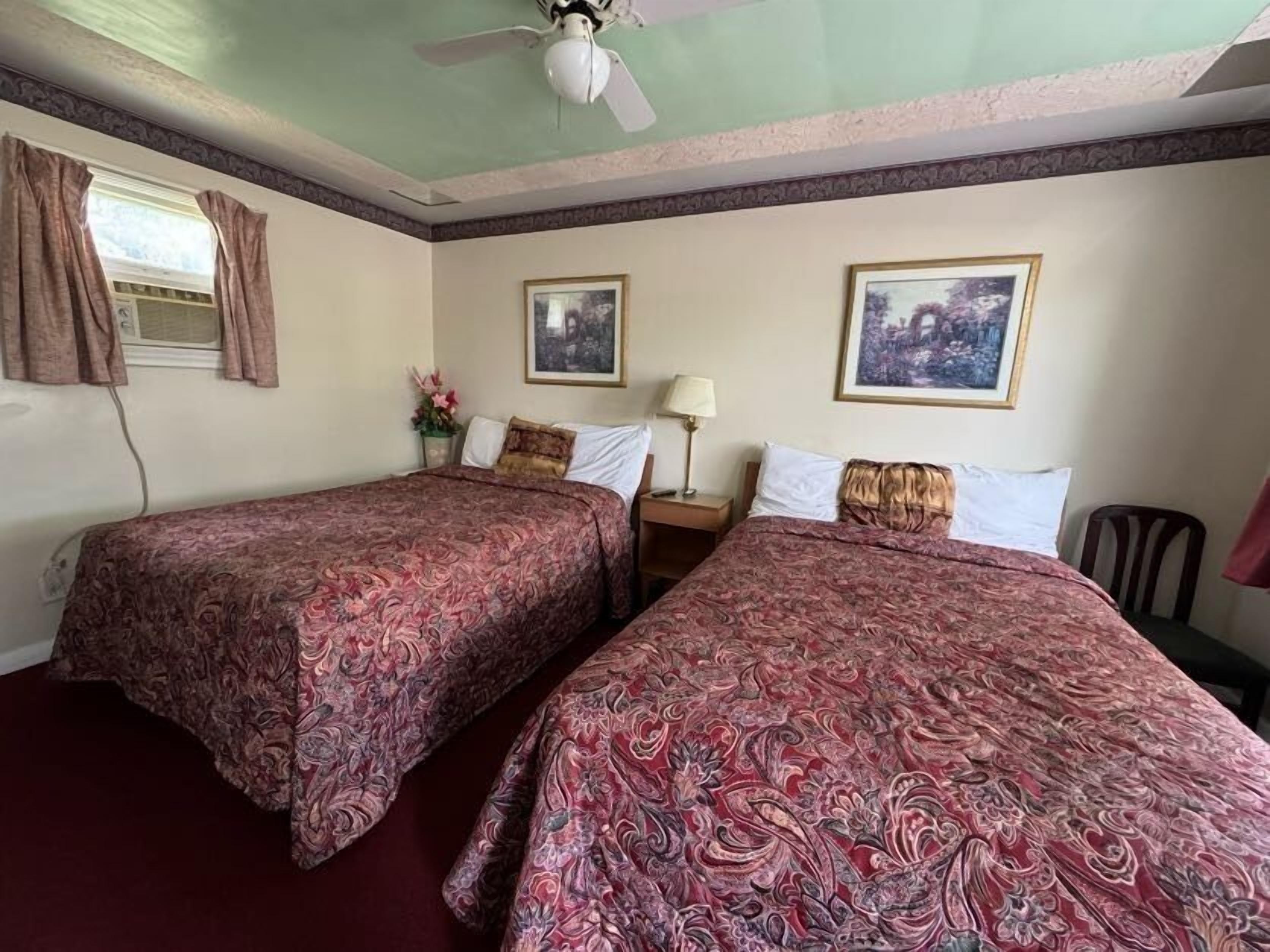 Habitación de hotel con dos camas, un ventilador de techo, una ventana con cortinas y dos cuadros en las paredes.