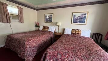 Habitación de hotel con dos camas, un ventilador de techo, una ventana con cortinas y dos cuadros en las paredes.