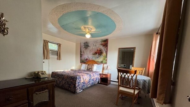 Habitación de hotel con cama, escritorio, silla, cómoda, ventilador de techo y un cuadro en la pared.