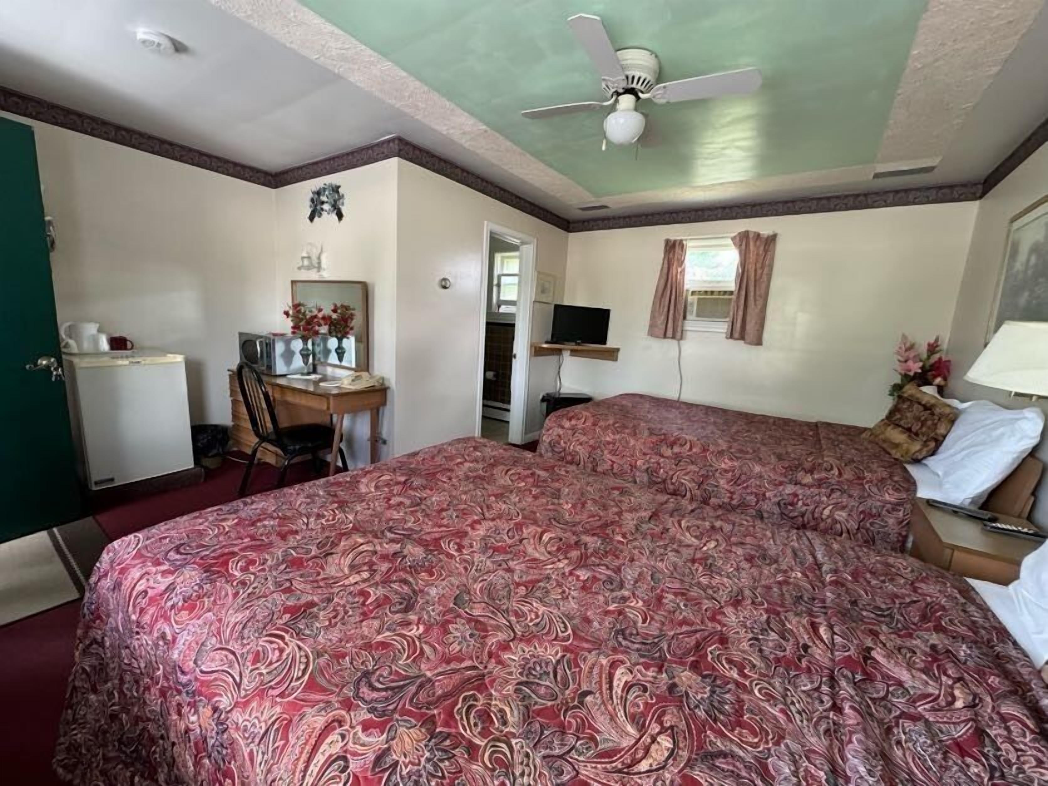 A hotel room with two beds, a desk with a chair, a television, and a ceiling fan.