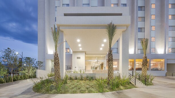 A modern hotel entrance with a covered walkway, palm trees, and a well-lit interior.
