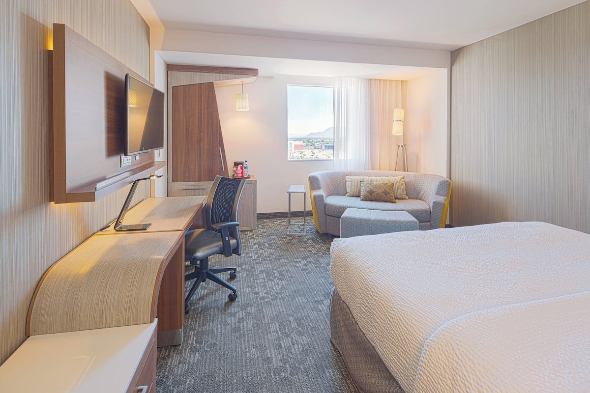 A hotel room with a bed, a desk with a chair, a TV, and a view of the city.